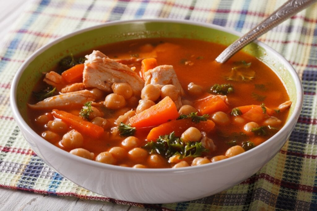 Sopa de grão-de-bico com frango, cenoura e coentro em um recipiente branco com uma colher de metal 