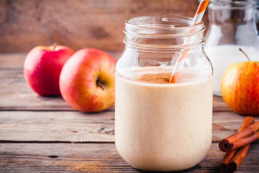 Copo de vidro com vitamina de maçã decorada com canela em pó em cima de uma mesa de madeira com maçãs