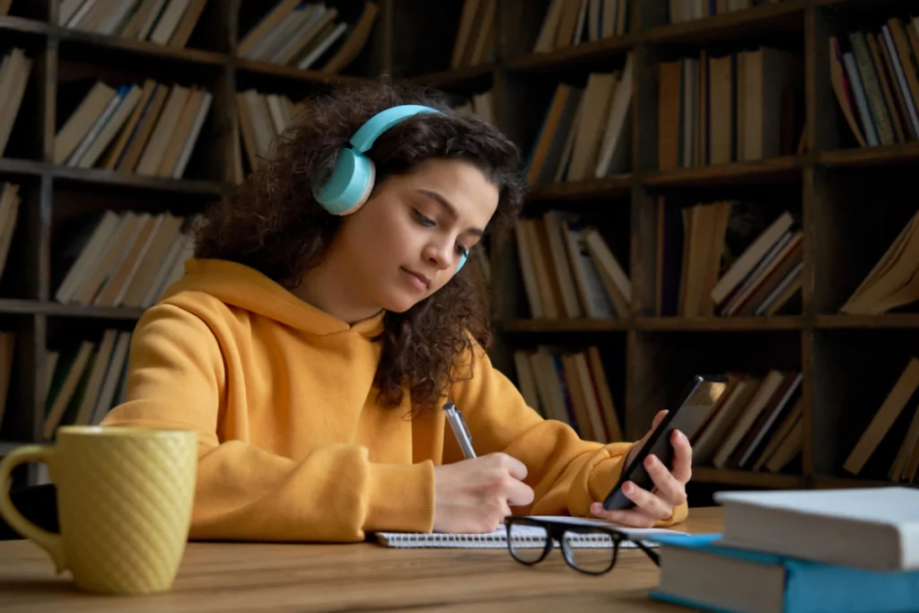 menina em ambiente com livro sentada escrevendo em caderno olhando o celular