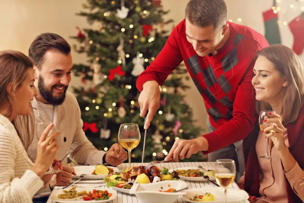 Pessoas sentadas em uma mesa de ceia de Natal e um homem em pé cortando pernil