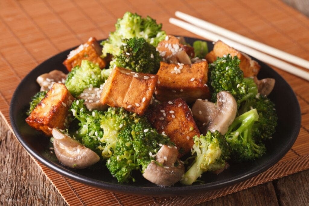 Brócolis com tofu e cogumelo servido em prato fundo preto em cima de mesa de madeira