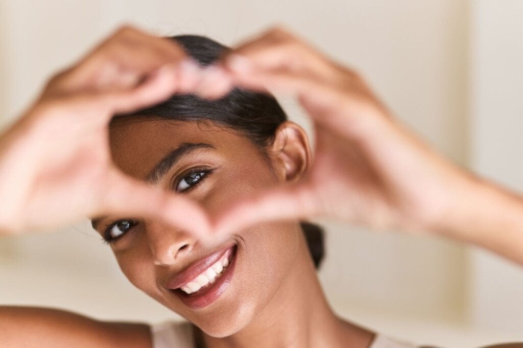 Mulher sorrindo fazendo um coração com as mãos em frente ao olho 