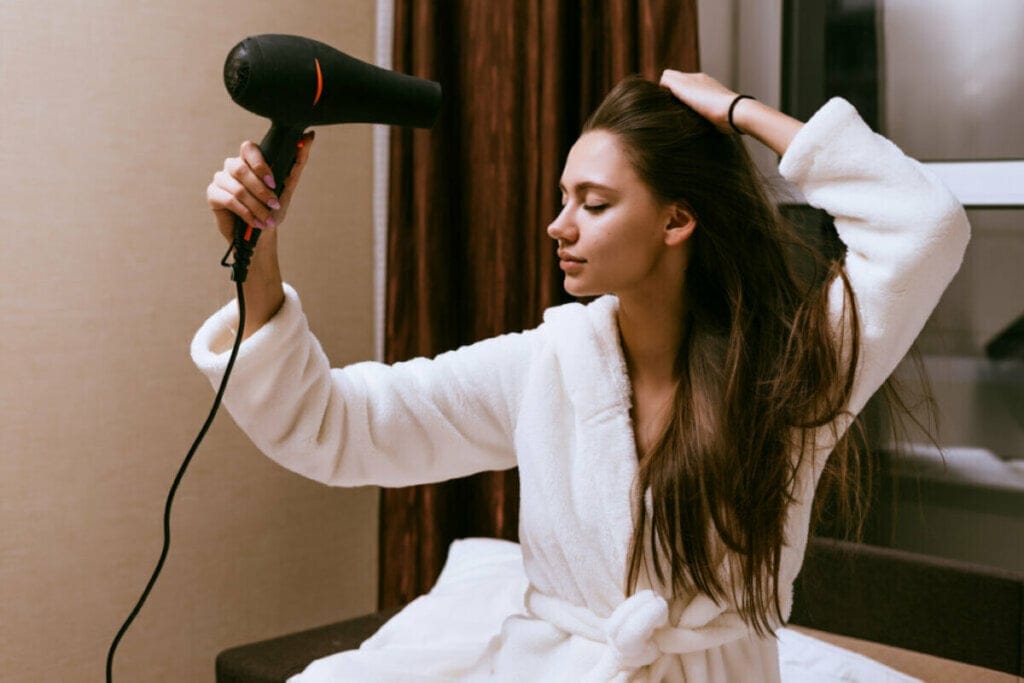 Mulher de roupão branco, sentada e secando o cabelo longo com um secador preto