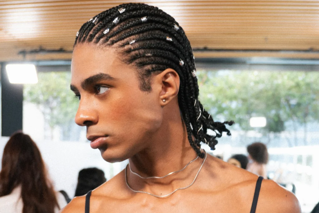 Homem com o cabelo trançado e com anéis pratas nas tranças usando uma blusa de alcinha preta