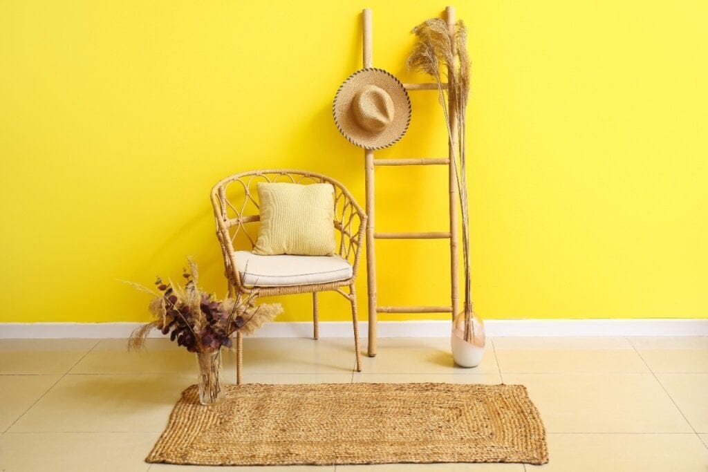 Ambiente com decoração tropical, parede amarela, cadeira de fibra natural com almofada, escada de bambu com chapéu e vaso com folhagens secas