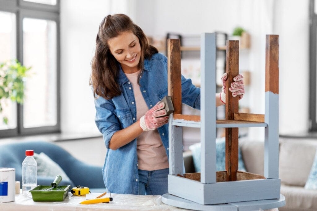 mulher de camisa jeans sorrindo enquanto pinta de azul banco de madeira