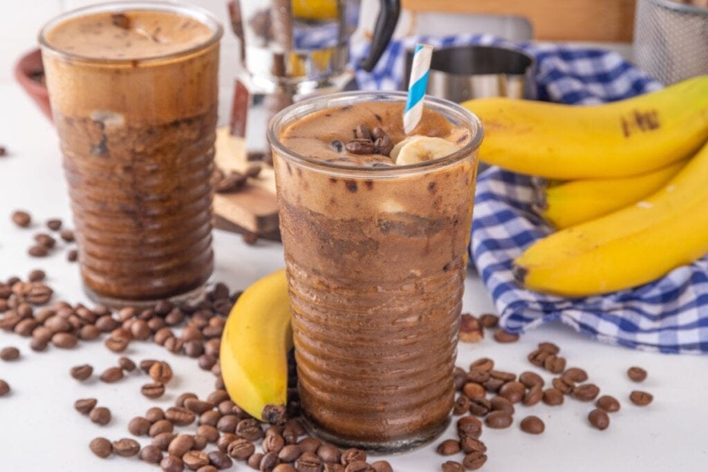 Smoothie de café e banana em dois copos de vidro sobre uma mesa com bananas e grãos de café