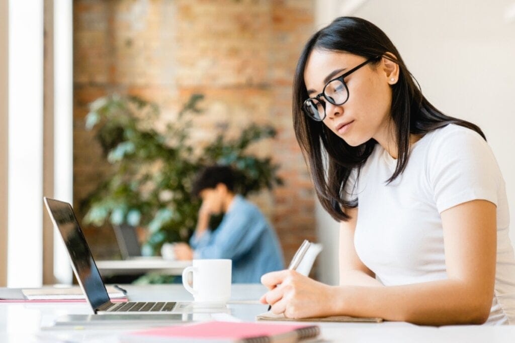 jovem de camiseta branca, sentada e escrevendo. notebook aberta à frente dela na mesa