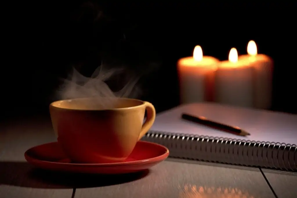 Xícara de café amarela ao lado de caderno e caneta e três velas ao fundo