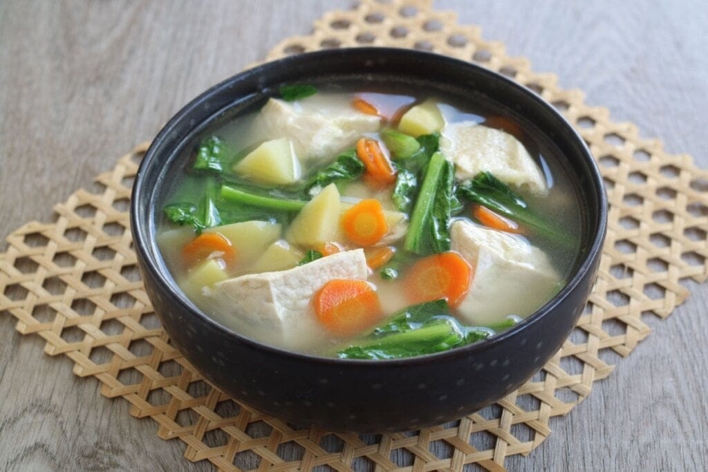 Sopa de tofu com cenoura, batata e espinafre servido em cumbuca de cerâmica preta em cima de descanso de panela em mesa de madeira