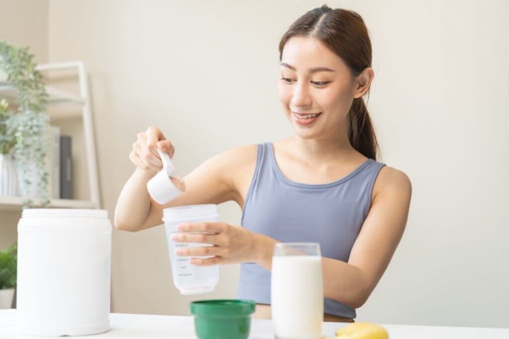 Jovem asiática com o cabelo preso em rabo de cavalo, usando uma regata roxa preparando shake de proteína 