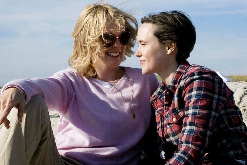 Duas mulheres sentadas juntas, uma com o cabelo curto loiro, usando óculos escuro e uma blusa rosa e uma calça bege e a outra tem o cabelo curto preto, usando uma camisa xadrez preta, vermelha e azul