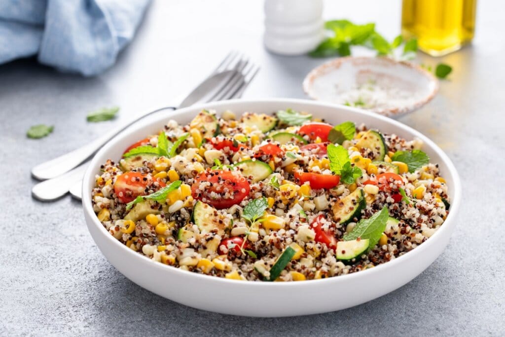 Recipiente branco com quinoa cozida, tomates-cereja, milho-verde, feijão-fradinho e folhas de hortelã em cima de uma mesa cinza 
