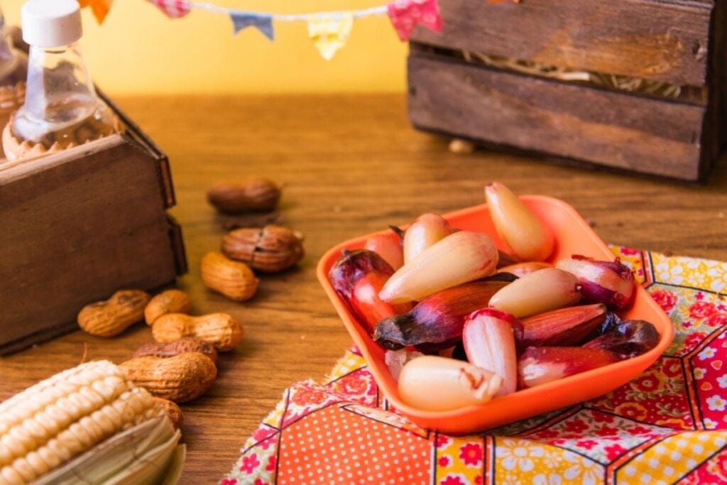 Prato com pinhão cozido, ao lado há uma espiga de milho e amendoim. Em uma mesa montada de festa junina