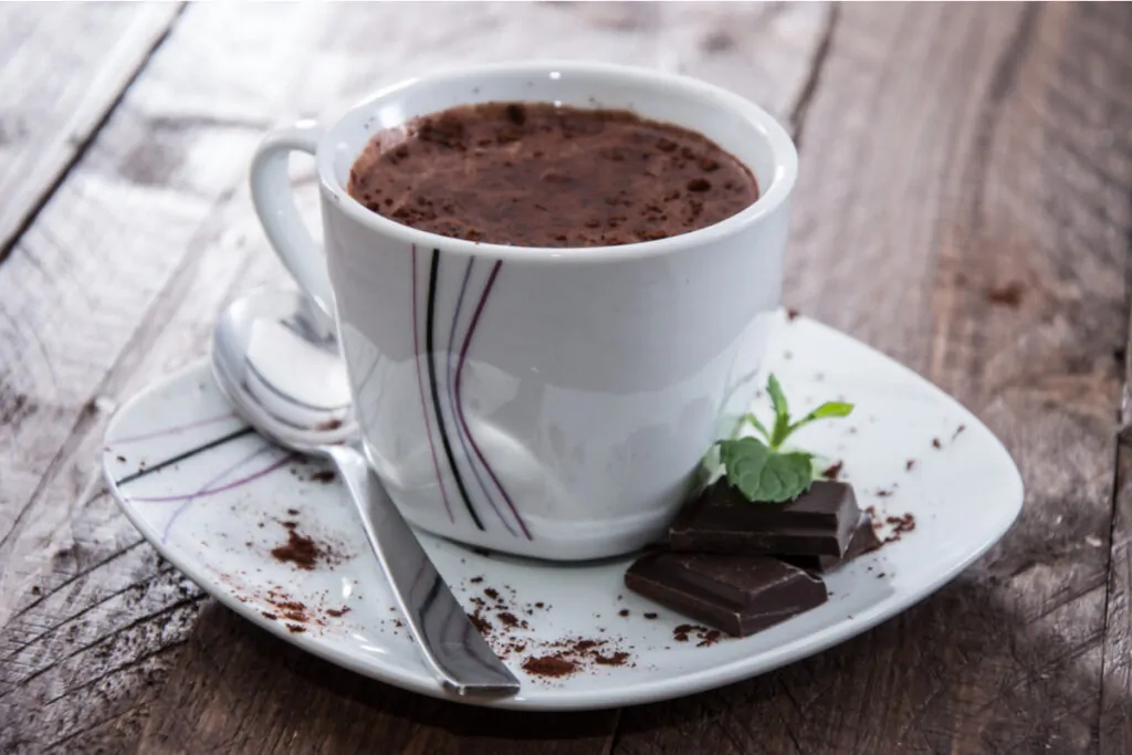 xícara branca em pires branco com chocolate quente com cacau em pó. dois tabletes de chocolate com hortelã no pires