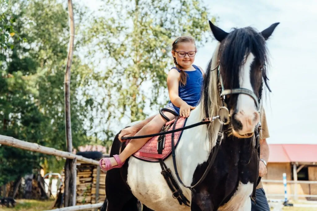 Menina com o cabelo preso em trança, usando regata azul, e short jeans e óculos andando a cavalo com ajuda de adulto