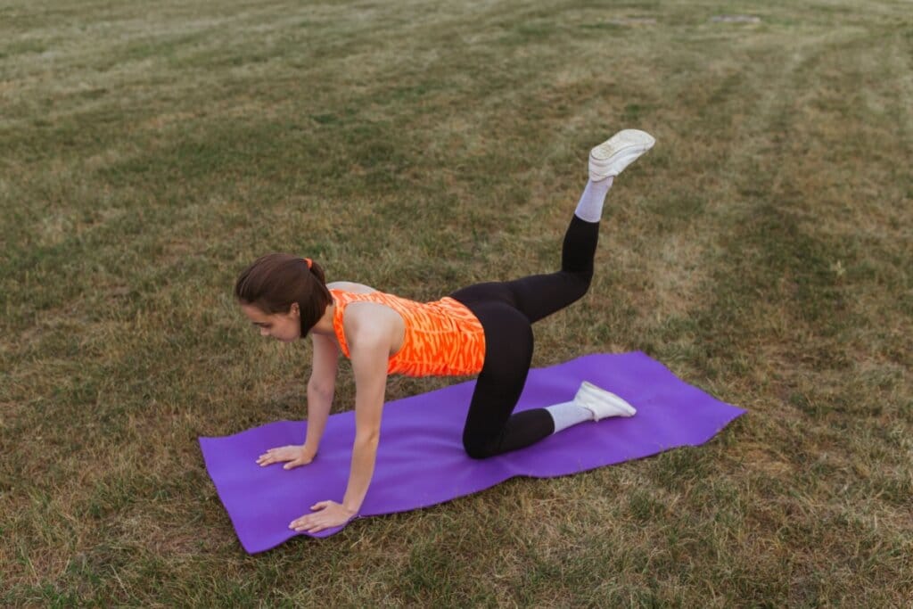 Mulher em quatro apoios em um tapete de ioga roxo, em um parque, fazendo exercício de glúteo de quatro apoios