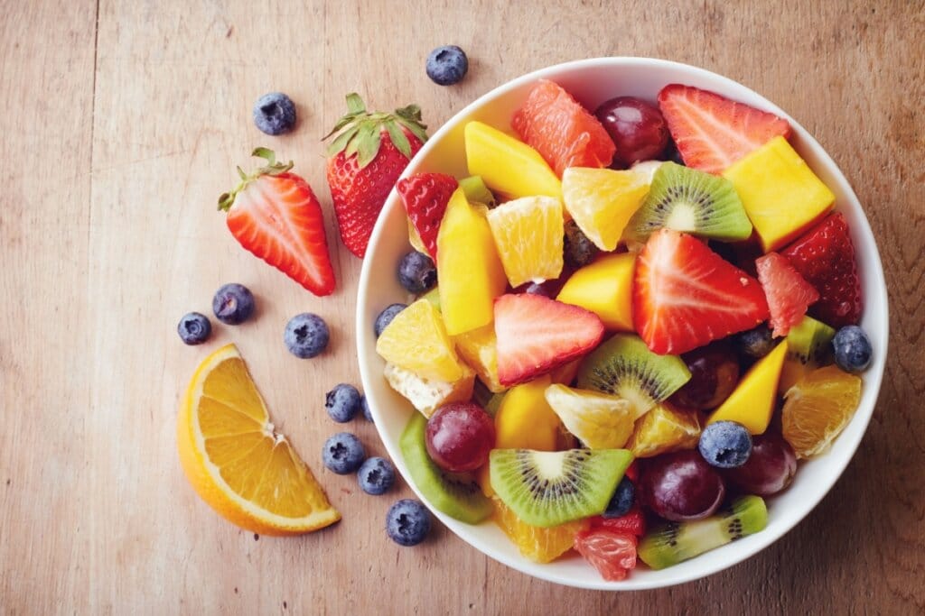 Recipiente branco com manga, kiwi, morango, mirtilo, uva e laranja em cima de uma mesa de madeira