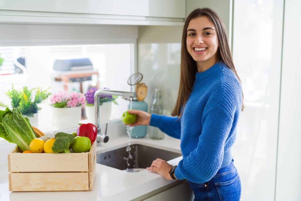 Mulher sorridente lavando maçã-verde em pia de cozinha, com caixa de frutas e verduras frescas ao lado, incluindo alface, brócolis e pimentão