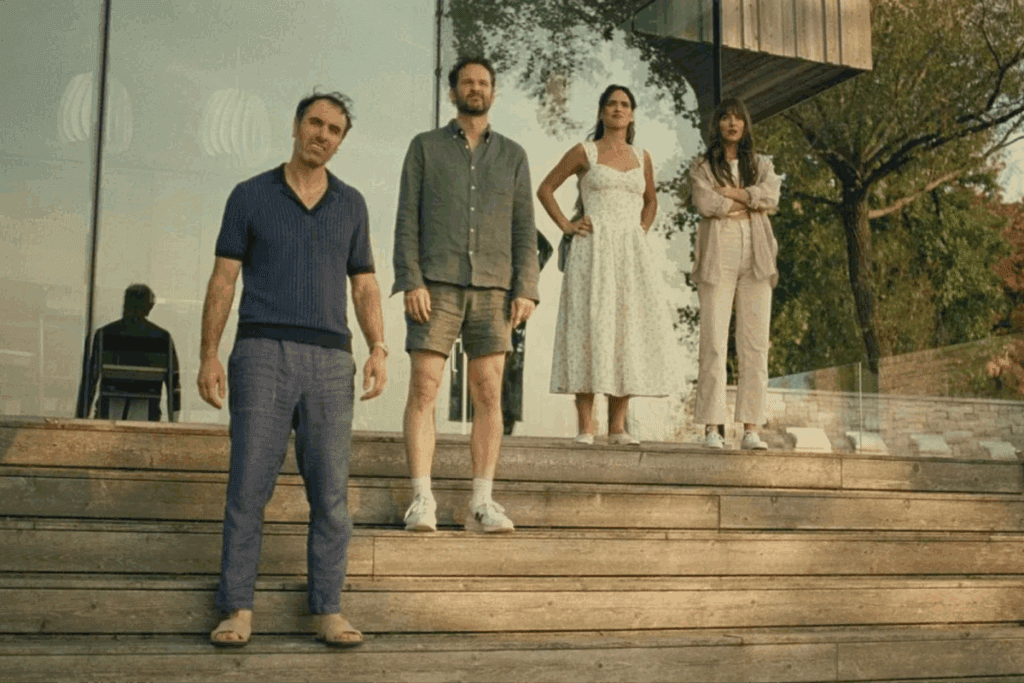 Quatro pessoas em pé sobre degraus de madeira em frente a uma casa de vidro, observando algo à frente no filme "Amores à Parte"