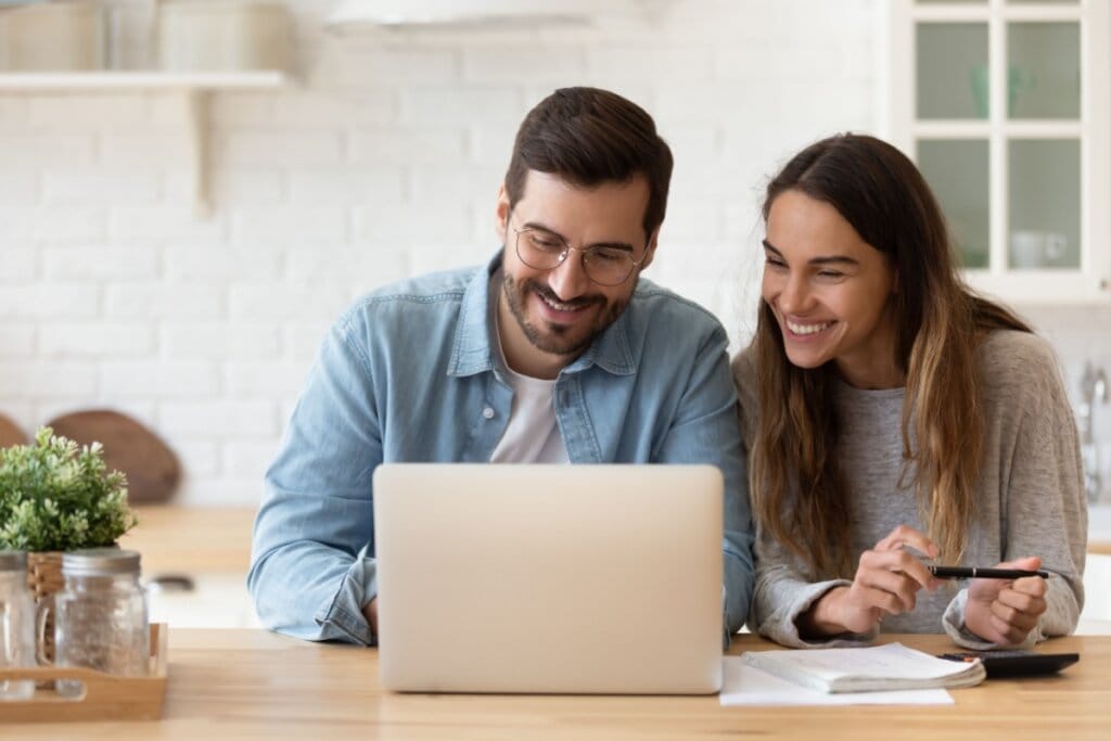 Casal sentado à mesa em frente ao notebook fazendo pesquisa. O homem tem o cabelo curto, usa óculos e barba, com uma camiseta branca e camisa jeans de botão e a mulher tem o cabelo longo, liso e solto. E está usando uma camisa de manga longa cinza
