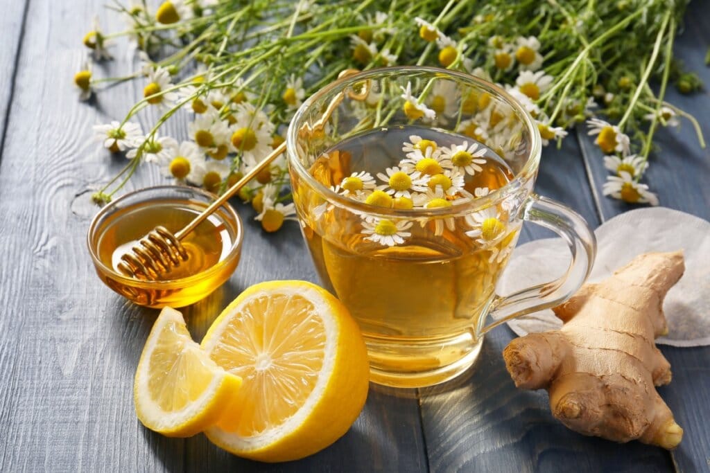 Chá de camomila com gengibre e limão-siciliano servido em xícara de vidro em cima de mesa de madeira com flores de camomila e gengibre e limão-siciliano