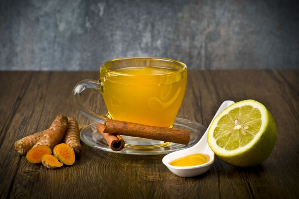 Chá de cúrcuma com canela e limão servido em xícara de vidro em cima de mesa de madeira com mel, limão e cúrcuma ao lado