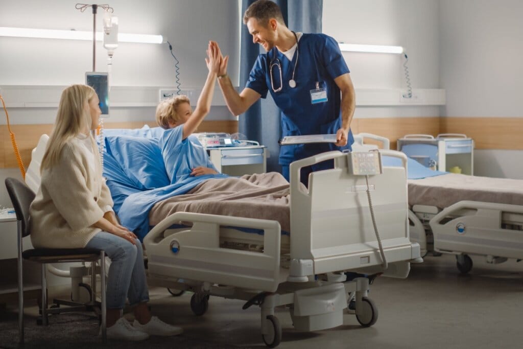 Menino em recuperação hospitalar após cirurgia, dando um high five ao médico enquanto a mãe acompanha ao lado do leito