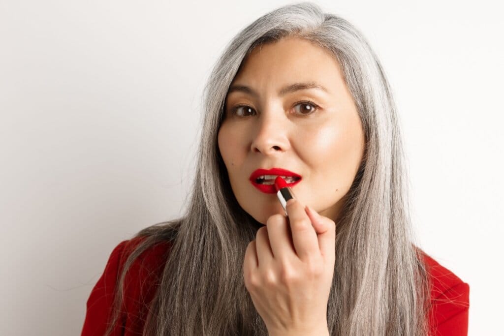 Mulher de cabelos grisalhos aplicando batom vermelho diante de fundo branco