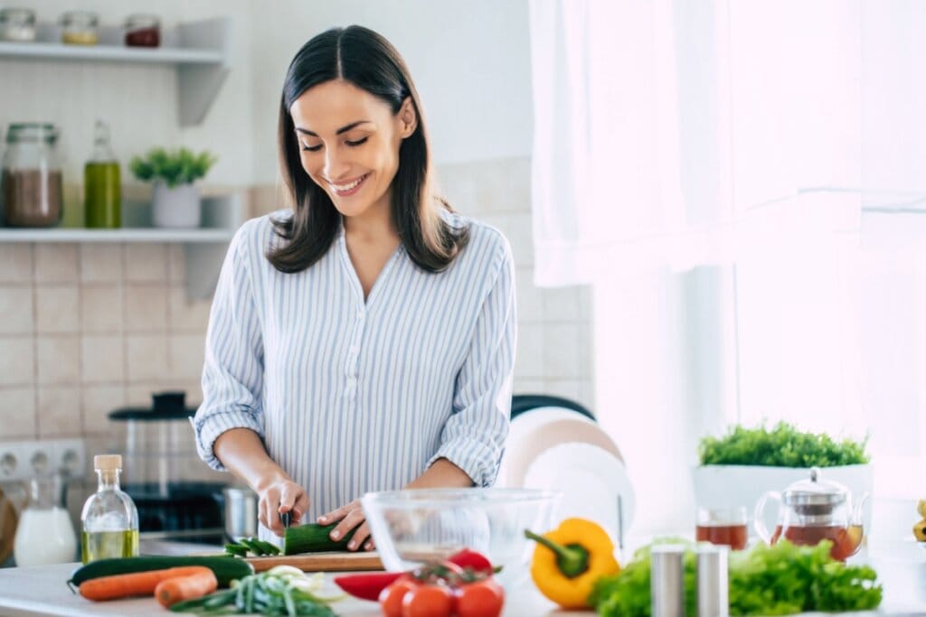 mulher de camisa branca e azul, sorrindo e picando legumes em tábua. à frente dela, há diversos alimentos vegetais