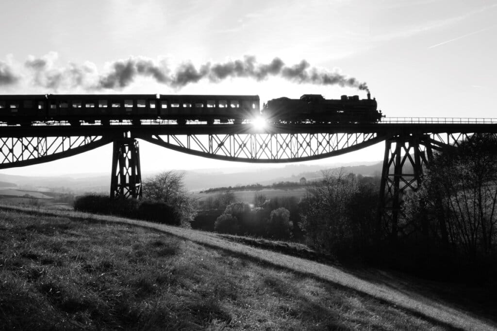 Imagem em preto e branco de um trem a vapor em trilhos no alto de uma ponte 