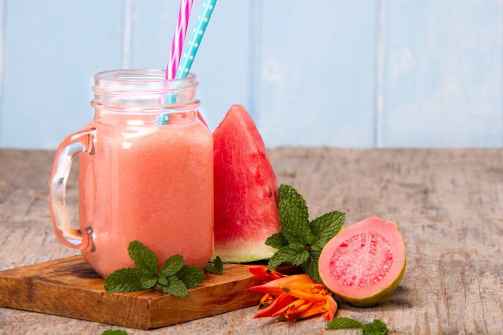 Suco de goiaba com melancia servido em caneca de vidro com folhas de hortelã ao lado, junto com goiaba cortada ao meio e fatia de melancia em cima de tábua de madeira em cima de mesa de madeira