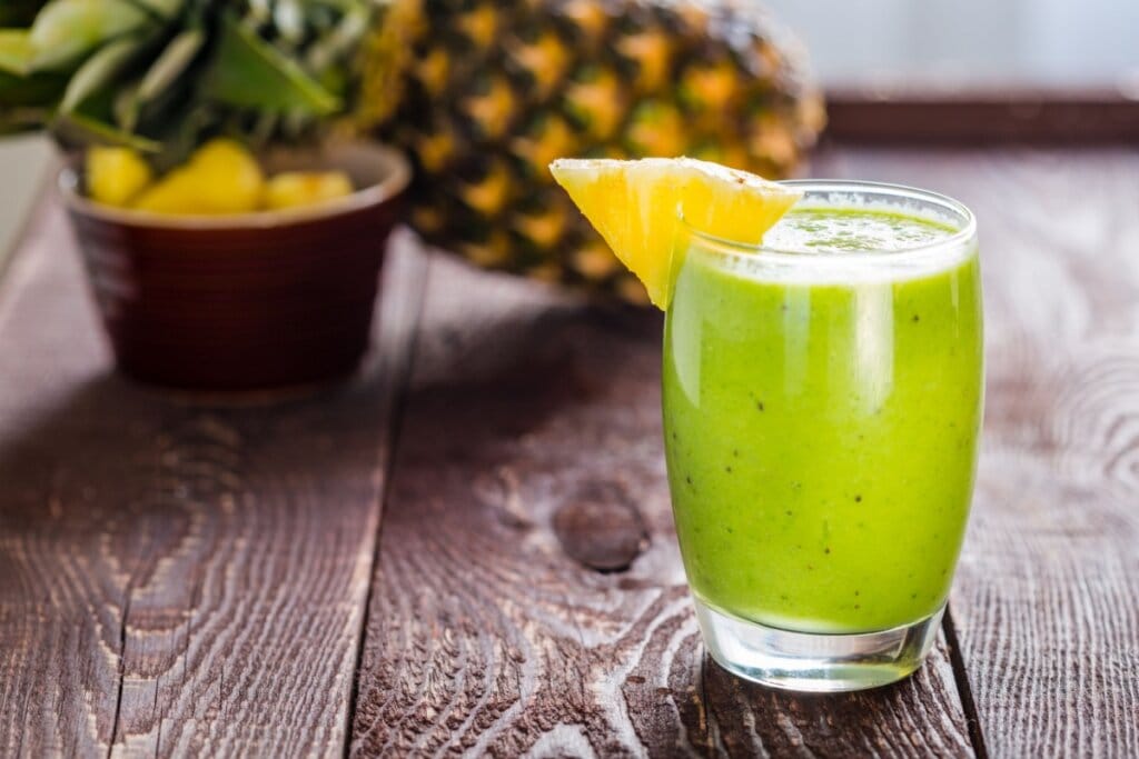 Suco de abacaxi com espinafre e couve em copo de vidro em superfície de madeira; ao fundo, recipiente com pedaços de abacaxi e abacaxi inteiro apoiado