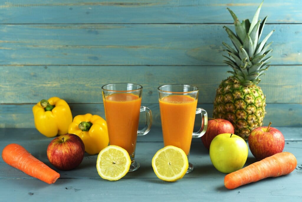 Suco detox de abacaxi, cenoura, maçã e pimentão amarelo em dois copos rodeados pelos ingredientes usados