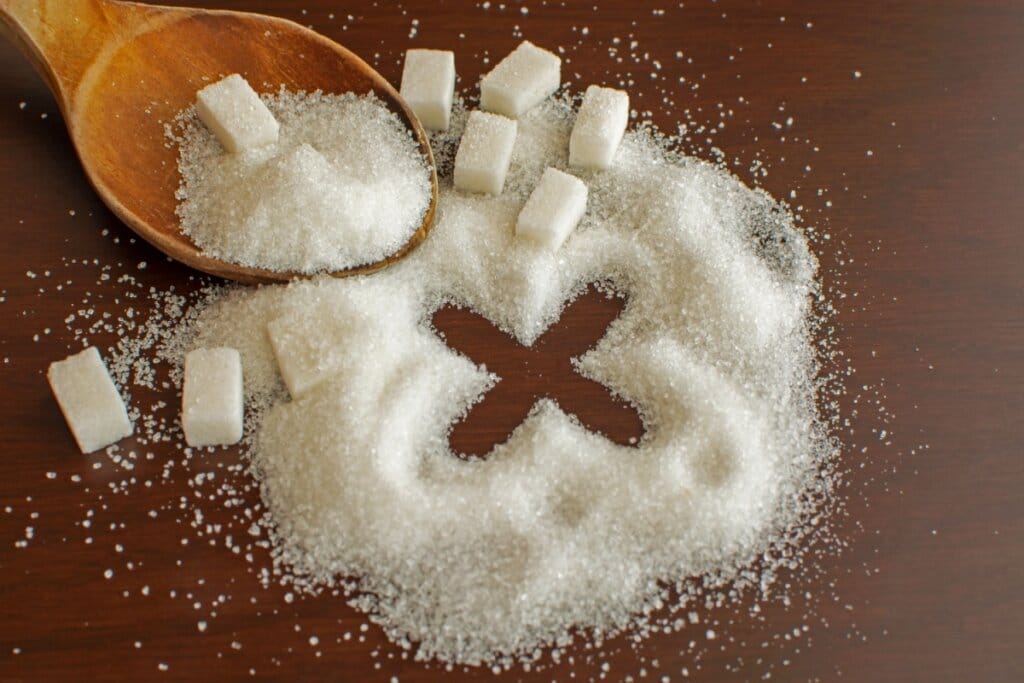 Açúcar esparramado em mesa de madeira com o desenho de um X e com diversos cubos de açúcar ao redor e uma colher de madeira com açúcar ao lado