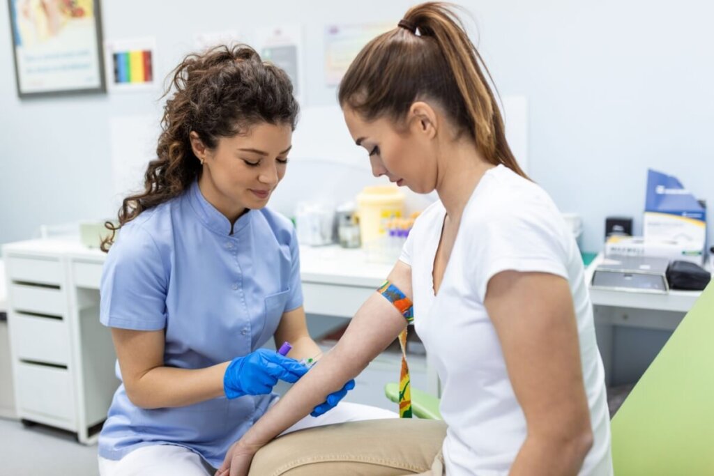 Mulher com cabelo amarrado em rabo de cavalo usando camiseta branca com o braço sendo preparado para exame de sangue, e enfermeira com o cabelo cacheado e usando jaleco azul fazendo a coleta