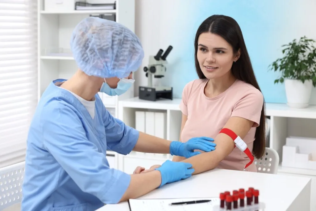 Profissional de saúde com máscara e touca preparando braço de paciente para coleta de sangue