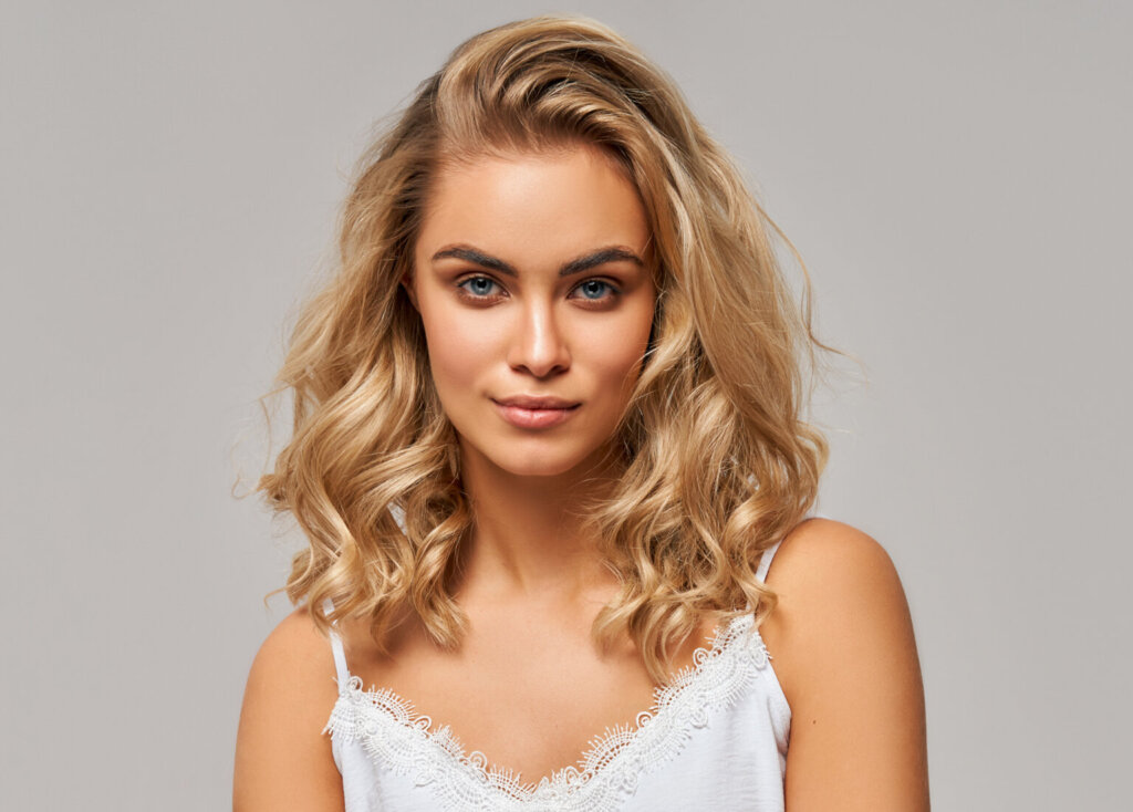 Mulher com cabelo loiro, ondulado até o ombro, usando regata branca em fundo cinza
