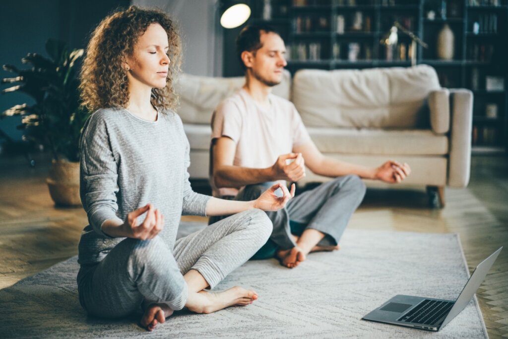 Homem e mulher meditando em posição de lótus na sala, com olhos fechados