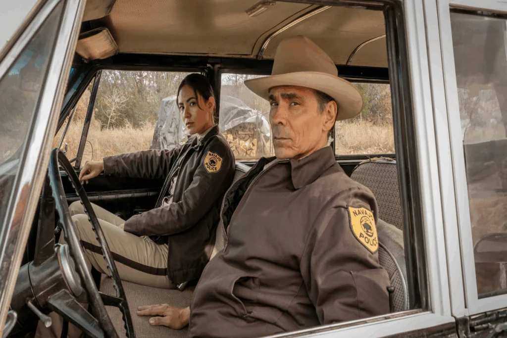 Imagem de dois policiais sentados dentro de uma viatura usando uniformes de polícia e o homem está usando um chapéu bege. 