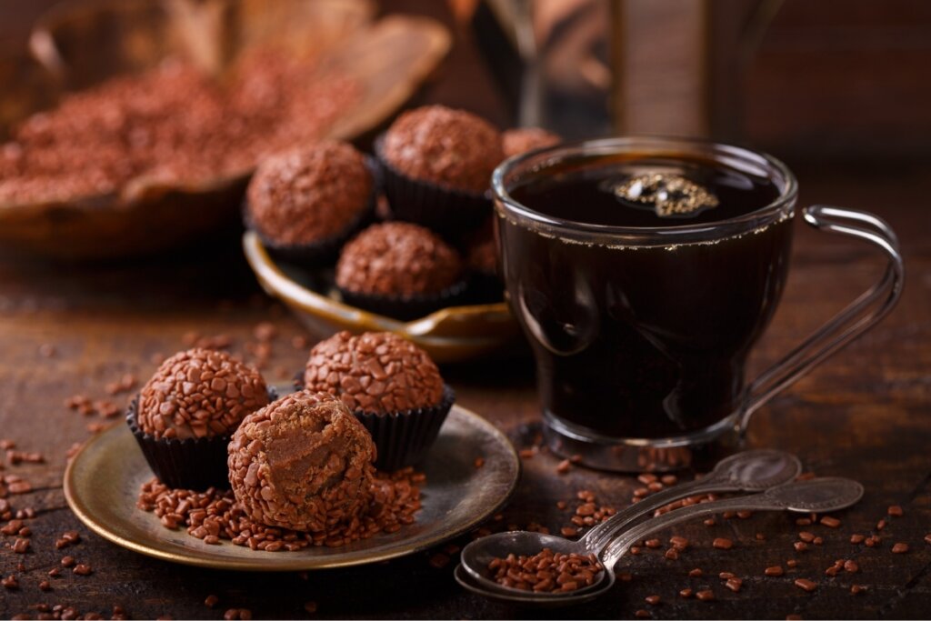 Três brigadeiros em cima de um prato com uma xícara com café preto ao lado