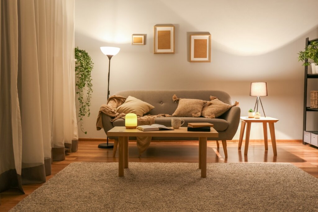 Sala de estar com iluminação amarela, sofá bege, mesa de centro e parede branca com três quadros ao fundo