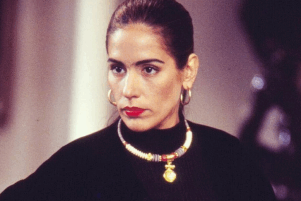 Mulher de expressão séria, cabelos presos e batom vermelho, usando blusa preta e colar dourado