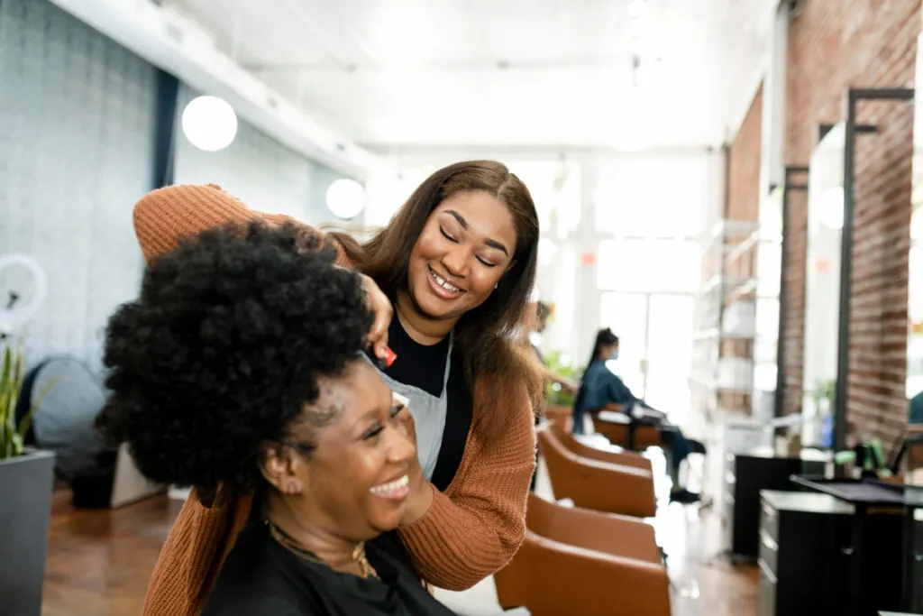 Cabeleireira de blusa marrom sorrindo e mexendo em cabelo de mulher de blusa preta sentada em cadeira em salão de beleza