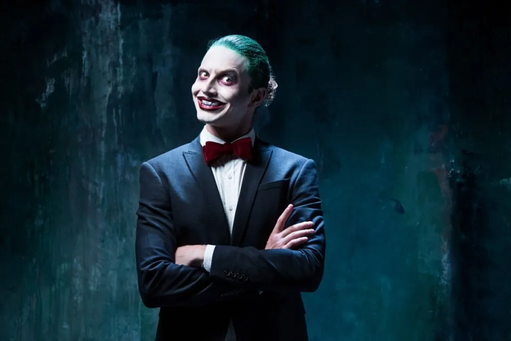 Homem caracterizado como Coringa, com cabelo verde, maquiagem marcante e terno elegante, exibindo um sorriso sinistro em um ambiente escuro