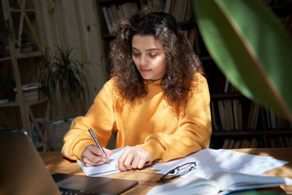 jovem de blusa amarela sentada e escrevendo em caderno. folhas e notebook em cima de mesa