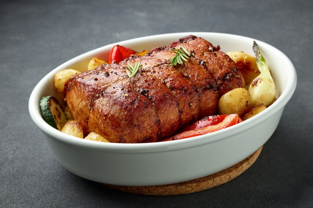 Lombo suíno amarrado com barbante culinário, assado e dourado, servido em travessa branca com batatas, abobrinha, tomate e cebola, decorado com alecrim fresco