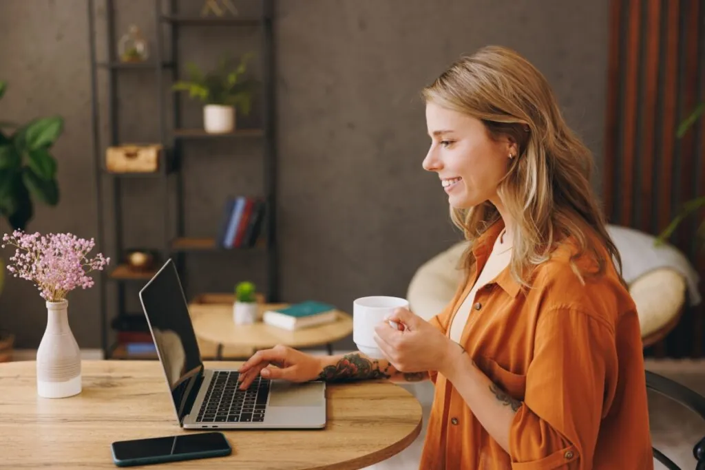 mulher loira, de blusa laranja, sentada, segurando xícara branca e digitando em notebook