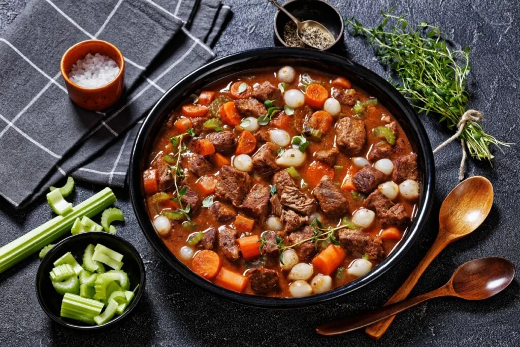 travessa preta com picadinho de carne com cenoura e minicebola. em volta, salsão, tomilho, colher de pau e sal grosso