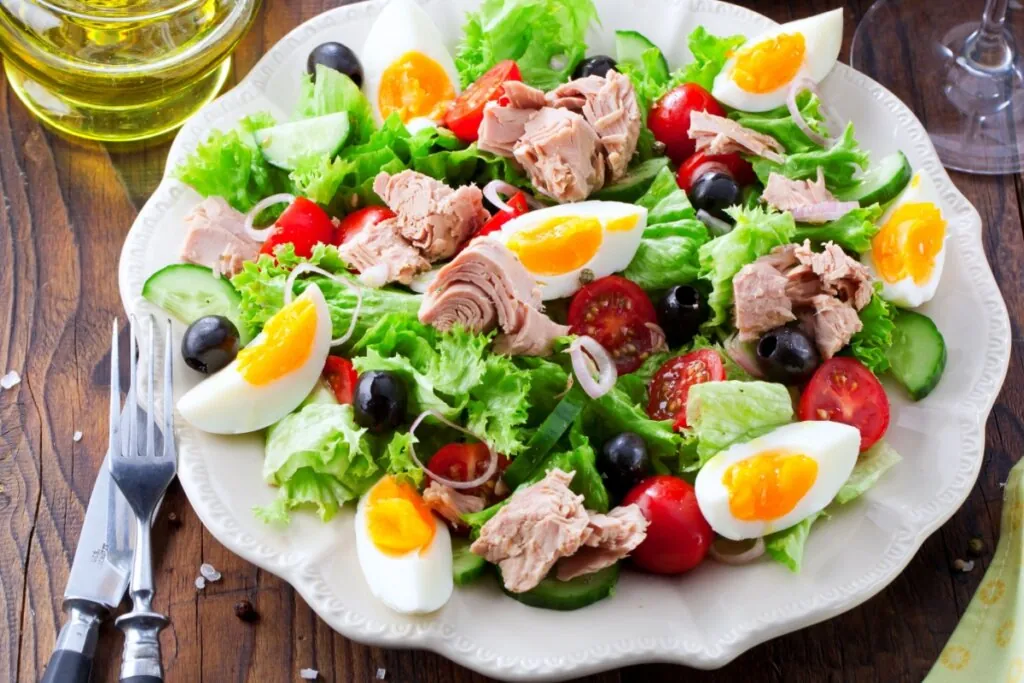 Salada de atum com ovos e legumes servido em prato branco em cima de mesa de madeira